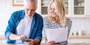 Older couple smiling while reviewing documents at a kitchen counter, with a smartphone and laptop nearby.
