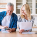 Older couple smiling while reviewing documents at a kitchen counter, with a smartphone and laptop nearby.