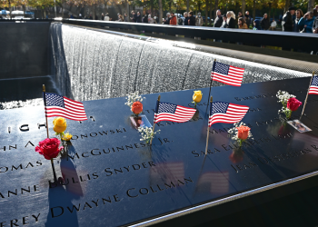 Small American flags and flowers rest on engraved panels beside a cascading memorial pool as visitors gather in the background.