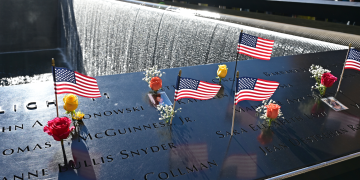Small American flags and flowers rest on engraved panels beside a cascading memorial pool as visitors gather in the background.
