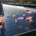 Small American flags and flowers rest on engraved panels beside a cascading memorial pool as visitors gather in the background.