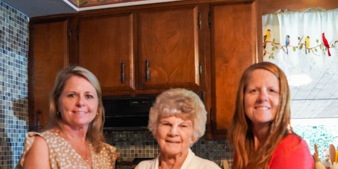 Three women pose in a kitchen while holding a homemade butterscotch pie with meringue.