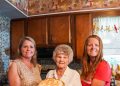 Three women pose in a kitchen while holding a homemade butterscotch pie with meringue.