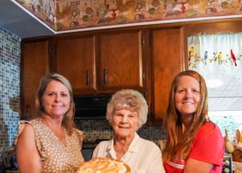 Three women pose in a kitchen while holding a homemade butterscotch pie with meringue.