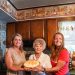 Three women pose in a kitchen while holding a homemade butterscotch pie with meringue.