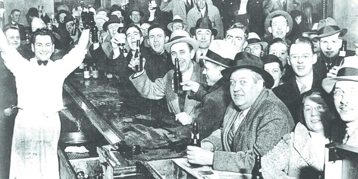 A crowd of men in a historic bar raise bottles and glasses in celebration after Prohibition ended.