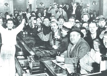 A crowd of men in a historic bar raise bottles and glasses in celebration after Prohibition ended.