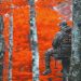 A hunter in camouflage sits in a tree stand among orange autumn leaves.