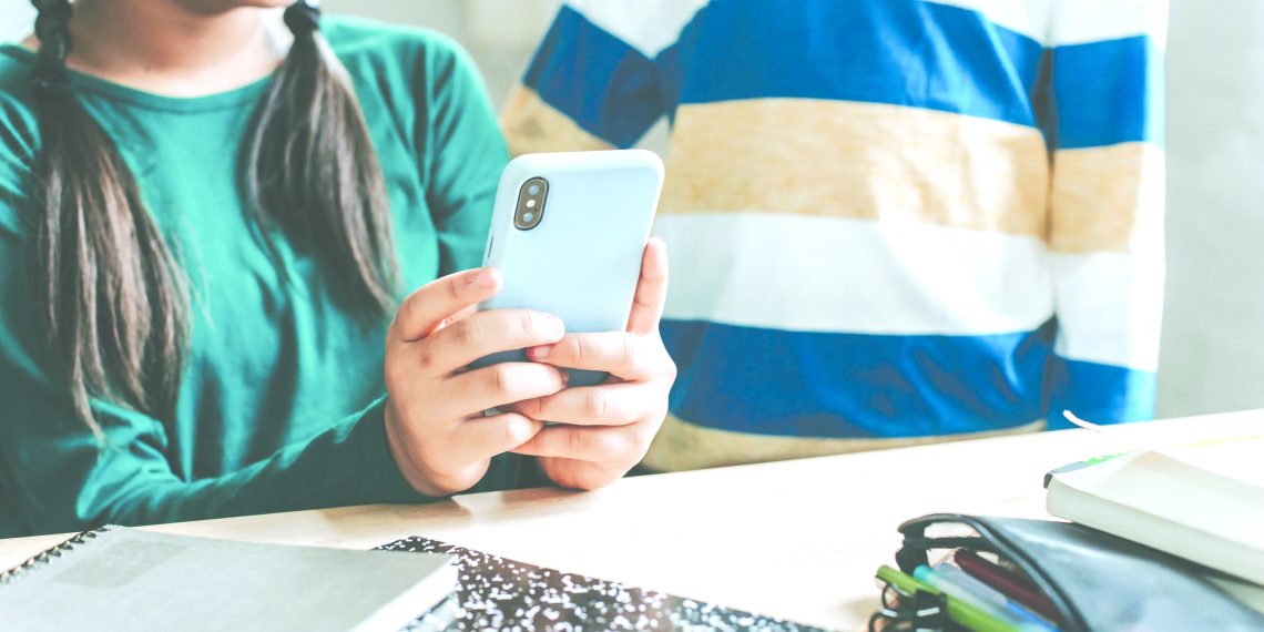 Two students in a classroom with one student holding a smartphone at a table with school supplies.