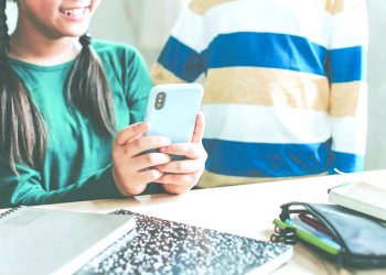 Two students in a classroom with one student holding a smartphone at a table with school supplies.