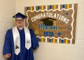 Mason smiles in a blue graduation cap and gown beside a handmade “Congratulations Mason” Class of 2026 sign.