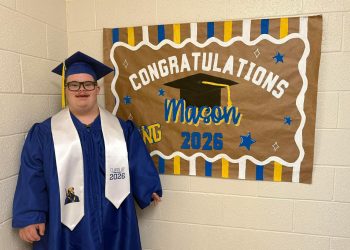 Mason smiles in a blue graduation cap and gown beside a handmade “Congratulations Mason” Class of 2026 sign.
