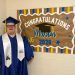 Mason smiles in a blue graduation cap and gown beside a handmade “Congratulations Mason” Class of 2026 sign.