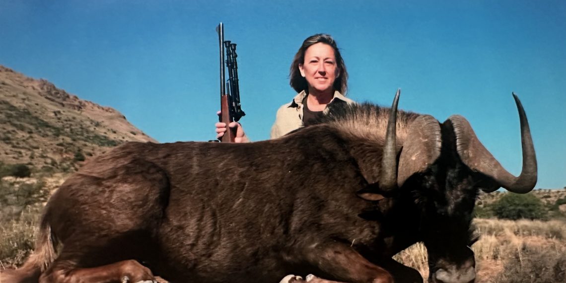 A woman holding a rifle stands behind a dead wild animal in a dry landscape under a blue sky.