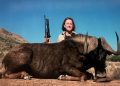 A woman holding a rifle stands behind a dead wild animal in a dry landscape under a blue sky.