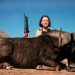 A woman holding a rifle stands behind a dead wild animal in a dry landscape under a blue sky.