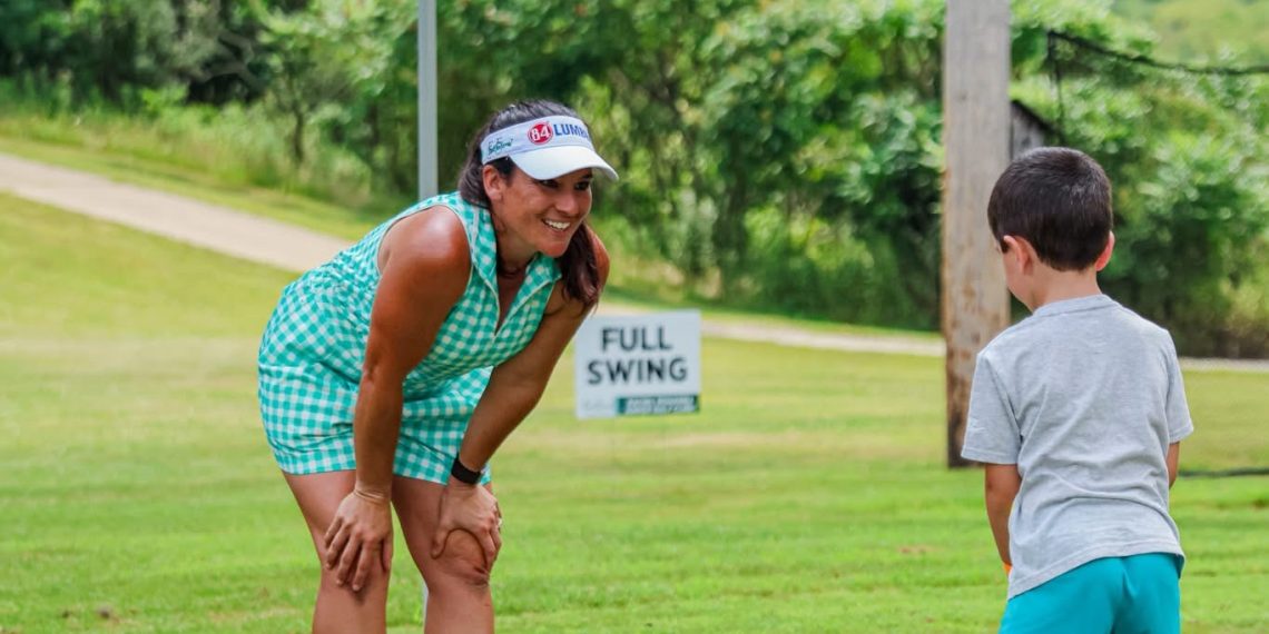 A woman in a turquoise golf outfit watches a young boy practice golf on a grassy course beneath a green First Federal canopy.