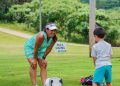 A woman in a turquoise golf outfit watches a young boy practice golf on a grassy course beneath a green First Federal canopy.