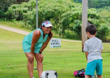 A woman in a turquoise golf outfit watches a young boy practice golf on a grassy course beneath a green First Federal canopy.