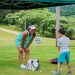 A woman in a turquoise golf outfit watches a young boy practice golf on a grassy course beneath a green First Federal canopy.