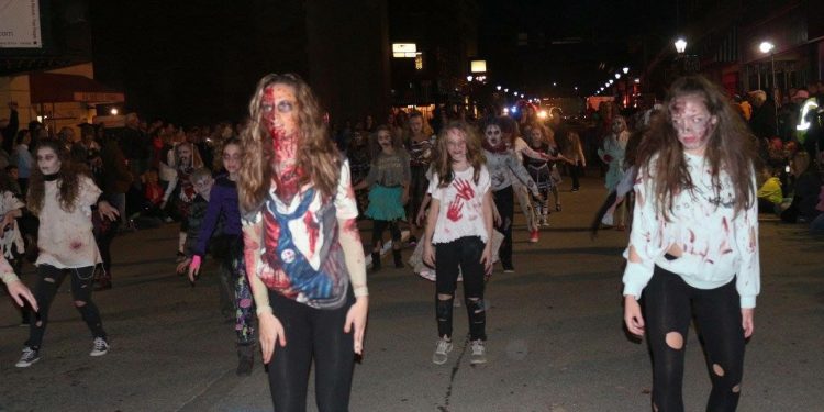 A large group of people dressed as zombies performs a Thriller-inspired dance on a downtown street at night.