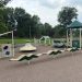 Accessible playground with colorful play equipment, a wavy balance feature, spinner, climbing structures, and shaded pavilion surrounded by trees and grass.