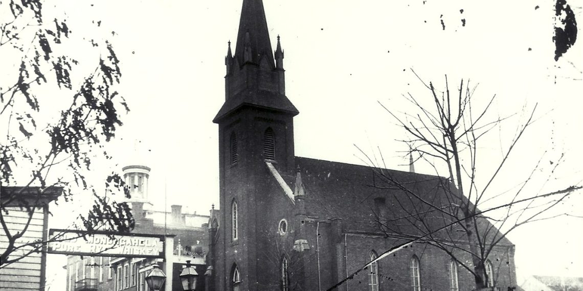 Historic black-and-white photograph of a Gothic church and downtown Waynesburg buildings, with a “Monongahela Whiskey” sign visible in the foreground.