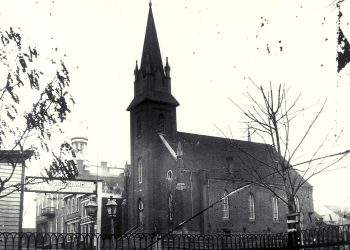 Historic black-and-white photograph of a Gothic church and downtown Waynesburg buildings, with a “Monongahela Whiskey” sign visible in the foreground.