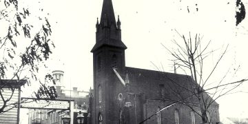 Historic black-and-white photograph of a Gothic church and downtown Waynesburg buildings, with a “Monongahela Whiskey” sign visible in the foreground.