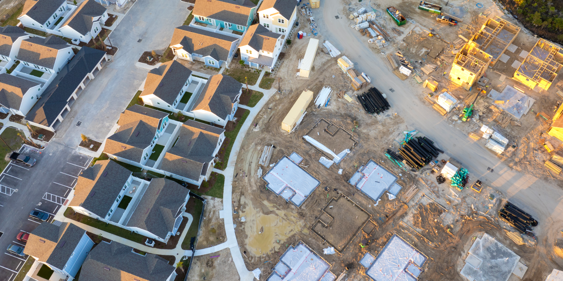 Aerial view of a residential neighborhood with completed homes beside an active construction site with foundations, building materials, and heavy equipment.