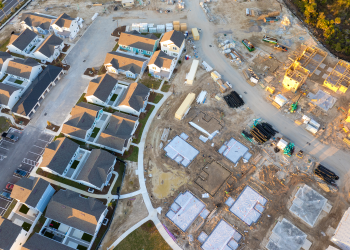 Aerial view of a residential neighborhood with completed homes beside an active construction site with foundations, building materials, and heavy equipment.