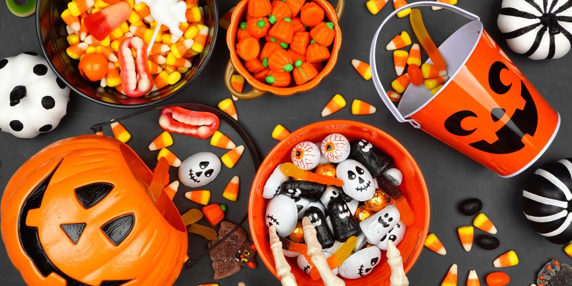 Halloween candy corn, chocolate and gummy candies in bowls and an orange jack-o’-lantern bucket surrounded by spooky decorations.