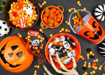 Halloween candy corn, chocolate and gummy candies in bowls and an orange jack-o’-lantern bucket surrounded by spooky decorations.