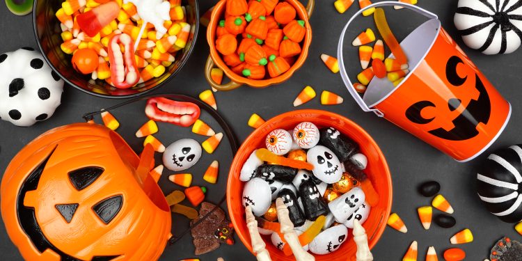 Halloween candy corn, chocolate and gummy candies in bowls and an orange jack-o’-lantern bucket surrounded by spooky decorations.