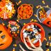 Halloween candy corn, chocolate and gummy candies in bowls and an orange jack-o’-lantern bucket surrounded by spooky decorations.