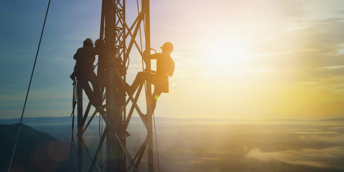 Two workers climb a tall metal utility tower at sunrise above a hazy landscape.
