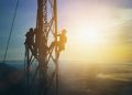 Two workers climb a tall metal utility tower at sunrise above a hazy landscape.