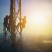 Two workers climb a tall metal utility tower at sunrise above a hazy landscape.