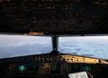 Airplane cockpit instruments and overhead controls with clouds and a snowy landscape visible through the windshield.