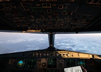 Airplane cockpit instruments and overhead controls with clouds and a snowy landscape visible through the windshield.