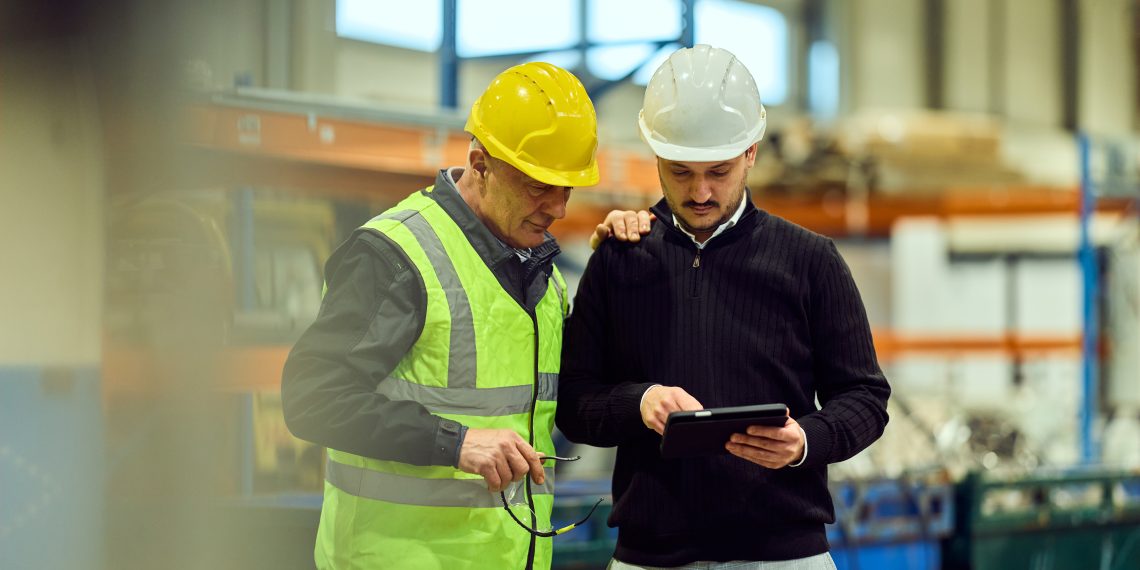 Two industrial workers in hard hats review a tablet together on a manufacturing floor.