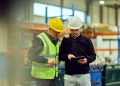 Two industrial workers in hard hats review a tablet together on a manufacturing floor.