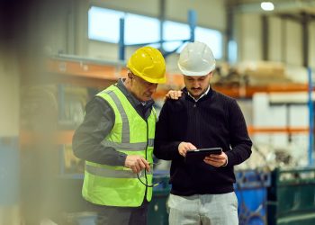 Two industrial workers in hard hats review a tablet together on a manufacturing floor.