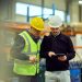 Two industrial workers in hard hats review a tablet together on a manufacturing floor.
