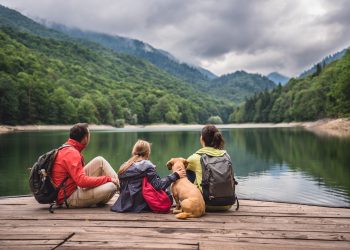 A family and their dog sit together on a wooden dock overlooking a peaceful mountain lake surrounded by green mountains.
