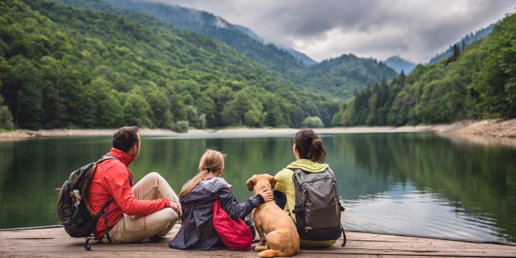 A family and their dog sit together on a wooden dock overlooking a peaceful mountain lake surrounded by green mountains.