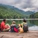 A family and their dog sit together on a wooden dock overlooking a peaceful mountain lake surrounded by green mountains.