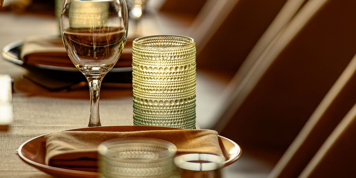 A warmly lit restaurant dining room with set tables, wine glasses, candles, and rows of upholstered chairs.