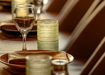 A warmly lit restaurant dining room with set tables, wine glasses, candles, and rows of upholstered chairs.