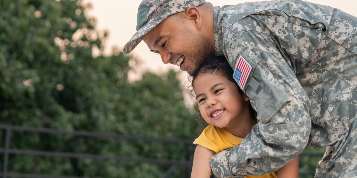 A smiling service member in camouflage hugs a smiling young child in a yellow shirt outdoors.